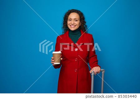 Charming delightful dark-haired woman in red coat smiles toothy smile looking at camera, holds a takeaway paper cup with hot coffee or tea and suitcase isolated over blue background with copy space Charming delightful dark-haired woman in red coat smiles toothy smile looking at camera, holds a takeaway paper cup with hot coffee or tea and suitcase isolated over blue background with copy space 85303440