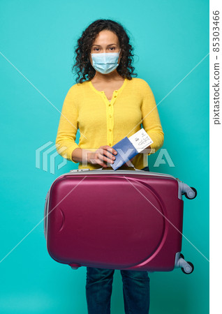 Young woman in protective medical mask, wearing bright yellow sweater posing with suitcase, passport and boarding pass in her hands. Travel, air flight journey concept on blue background, copy space Young woman in protective medical mask, wearing bright yellow sweater posing with suitcase, passport and boarding pass in her hands. Travel, air flight journey concept on blue background, copy space 85303466