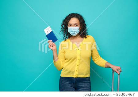 Cheerful Caucasian woman with curly dark hair, wearing protective medical mask, poses with a boarding pass and air ticket in the passport and a suitcase. Concept of safety travel during pandemic Cheerful Caucasian woman with curly dark hair, wearing protective medical mask, poses with a boarding pass and air ticket in the passport and a suitcase. Concept of safety travel during pandemic 85303467