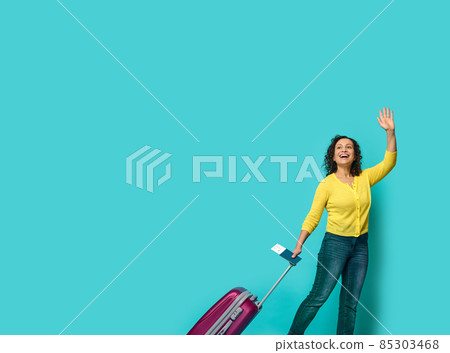 Full length of beautiful woman in casual clothes, waving hand, holding identity document and boarding pass, posing with suitcase, smiling looking to the side at a blue background with copy space Full length of beautiful woman in casual clothes, waving hand, holding identity document and boarding pass, posing with suitcase, smiling looking to the side at a blue background with copy space 85303468