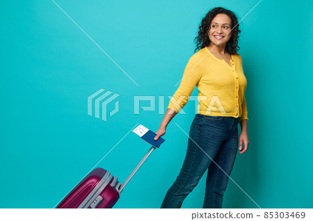 Happy curly dark-haired woman, traveler in bright yellow sweater and blue jeans holding passport with boarding pass and air ticket, smiles, posing with luggage on blue background with copy space Happy curly dark-haired woman, traveler in bright yellow sweater and blue jeans holding passport with boarding pass and air ticket, smiles, posing with luggage on blue background with copy space 85303469