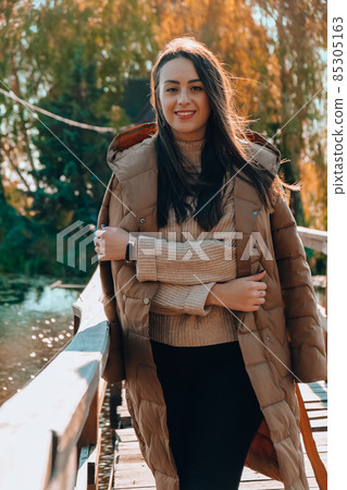 Beautiful brunette girl in autumn Park. Smiling young millennial woman in coat enjoying sunny weather in autumn fall weather. Outdoor portrait 85305163
