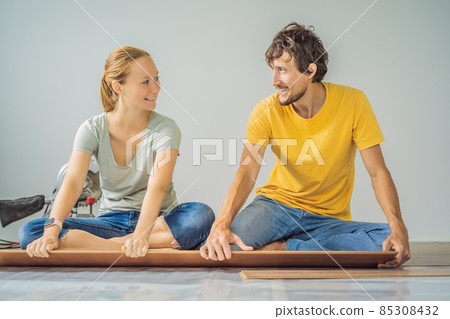 Married couple installing new wooden laminate flooring on a warm film floor. Infrared floor heating system under laminate floor Married couple installing new wooden laminate flooring on a warm film floor. Infrared floor heating system under laminate floor 85308432