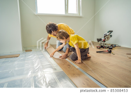 Father and son installing new wooden laminate flooring on a warm film floor. Infrared floor heating system under laminate floor 85308453