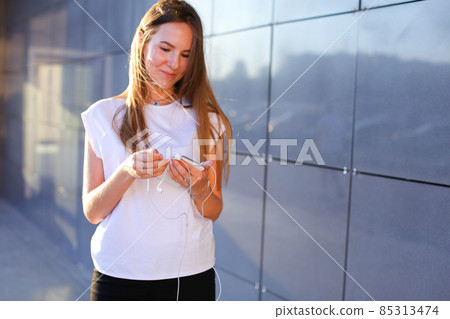 One young female woman unravels headphones for listening to music, talk on Skype, holds in hands phone, smiles and stands near wall of business center. One young female woman unravels headphones for listening to music, talk on Skype, holds in hands phone, smiles and stands near wall of business center. 85313474