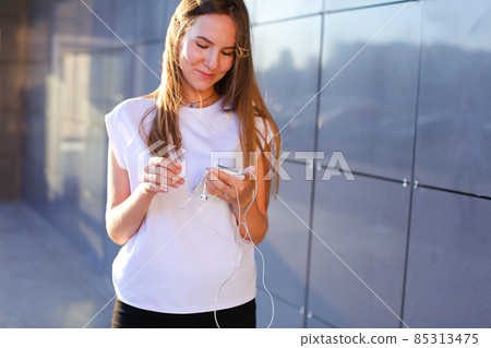 One young female woman unravels headphones for listening to music, talk on Skype, holds in hands phone, smiles and stands near wall of business center. One young female woman unravels headphones for listening to music, talk on Skype, holds in hands phone, smiles and stands near wall of business center. 85313475