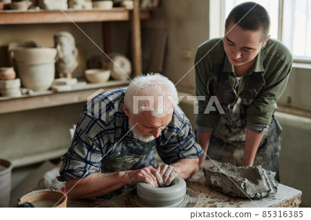 Senior potter sculpting a cup from clay pot with his assistant watching for the process in studio Senior potter sculpting a cup from clay pot with his assistant watching for the process in studio 85316385