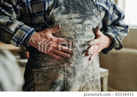 Close-up of a potter standing in a dirty apron and wiping his hands in clay on it while working in a pottery workshop 85316431