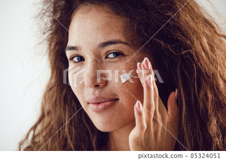 Portrait of woman with curly hair and freckles applying moisturizer to her face. 85324051