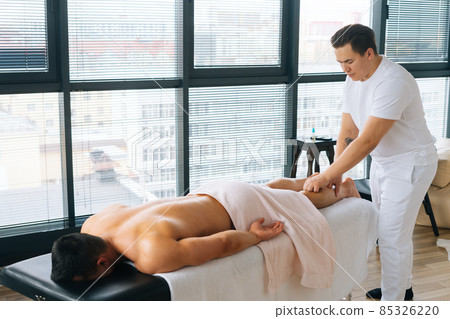 Professional male masseur with strong tattooed hands massaging lower leg of muscular athlete man after physical sports workout Professional male masseur with strong tattooed hands massaging lower leg of muscular athlete man after physical sports workout 85326220