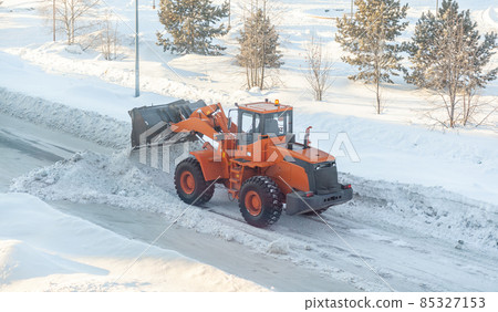 Big orange tractor cleans up snow from the road and loads it into the truck. Cleaning and cleaning of roads in the city from snow in winter Big orange tractor cleans up snow from the road and loads it into the truck. Cleaning and cleaning of roads in the city from snow in winter 85327153