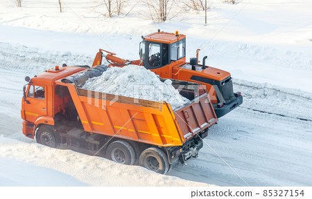 Big orange tractor cleans up snow from the road and loads it into the truck. Cleaning and cleaning of roads in the city from snow in winter 85327154