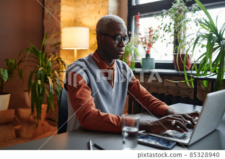 Serious African businessman in eyeglasses sitting at the table and typing on laptop working at office 85328940