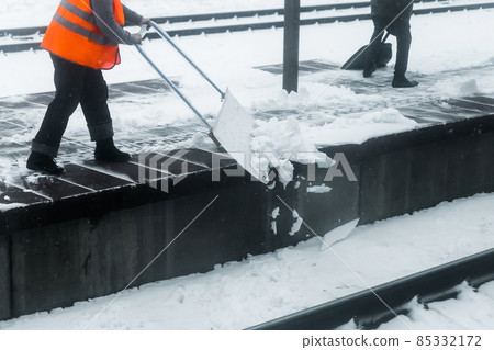 Woman worker in reflective orange vest cleans snow with a special shovel at the railway station Woman worker in reflective orange vest cleans snow with a special shovel at the railway station 85332172