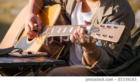 Man playing guitar at sunset lake, Close-up male hand playing on acoustic guitar outdoor with sunlight reflected on the water surface. 85339165