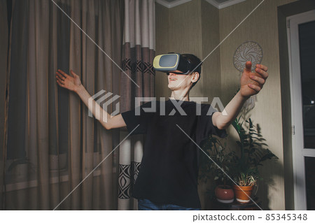 Young man wearing VR glasses met a friend in virtual reality and open his hands in greeting in the home interior. Communication in virtual reality. 85345348