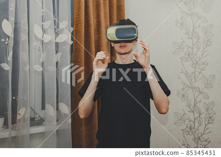 Young man in a VR helmet interacts with objects in virtual reality in the home interior. 85345431