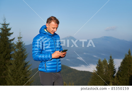 Male traveler using smartphone and smiling while standing outdoors with blue sky and mountains on background. Concept of hiking, travelling and mobile communication. Male traveler using smartphone and smiling while standing outdoors with blue sky and mountains on background. Concept of hiking, travelling and mobile communication. 85350159