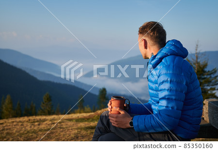 Hiking in the mountains. Guy sitting outdoors on mountain meadow at sunny morning, looking at very beautiful misty mountain hills. Active trekking recreation. Hiking in the mountains. Guy sitting outdoors on mountain meadow at sunny morning, looking at very beautiful misty mountain hills. Active trekking recreation. 85350160