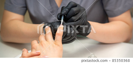 Painting female nails. Hands of manicurist in black gloves is applying transparent nail polish on female nails in a manicure salon. Painting female nails. Hands of manicurist in black gloves is applying transparent nail polish on female nails in a manicure salon. 85350368