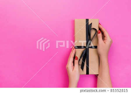Woman hands give wrapped valentine or other holiday handmade present in paper with black ribbon. Present box, decoration of gift on pink table, top view with copy space Woman hands give wrapped valentine or other holiday handmade present in paper with black ribbon. Present box, decoration of gift on pink table, top view with copy space 85352815