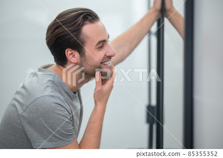 A young man in grey tshirt looking at his reflection in the mirror A young man in grey tshirt looking at his reflection in the mirror 85355352