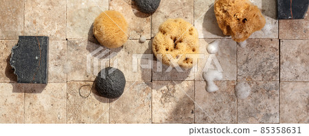 Bamboo charcoal bathroom accessories - konjac bamboo charcoal black sponge, handmade black soap and natural sea sponges with soap foam on earth toned stone tales top view 85358631
