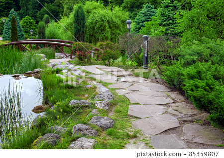 natural stone path along pond with water and reeds among green backyard plants with evergreen bushes and pine trees landscape design of eco friendly summer park with lantern, nobody. natural stone path along pond with water and reeds among green backyard plants with evergreen bushes and pine trees landscape design of eco friendly summer park with lantern, nobody. 85359807