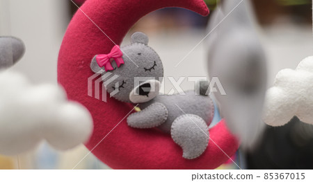 Baby crib mobile ornaments hangings close up shot, grey wool felt bear sleeps on a red plushie moon, surrounded by soft clouds. Baby crib mobile ornaments hangings close up shot, grey wool felt bear sleeps on a red plushie moon, surrounded by soft clouds. 85367015