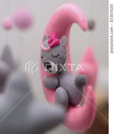 Baby crib mobile ornaments hangings close up, grey wool felt bear sleeps on a pink plushie moon, surrounded by stars. Baby crib mobile ornaments hangings close up, grey wool felt bear sleeps on a pink plushie moon, surrounded by stars. 85367020