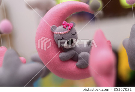 Baby crib mobile ornaments hangings close up, grey wool felt bear sleeps on a pink plushie moon, surrounded by stars. Baby crib mobile ornaments hangings close up, grey wool felt bear sleeps on a pink plushie moon, surrounded by stars. 85367021