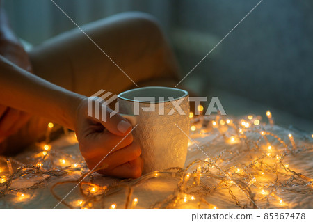 Womans hands holding a cup of tea in bed. Christmas lights on, cozy home atmosphere. Womans hands holding a cup of tea in bed. Christmas lights on, cozy home atmosphere. 85367478