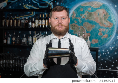A bearded adult man of European appearance, a professional bartender, holds in his hands in black latex gloves a tool for preparing and mixing alcoholic cocktails in a nightclub 85368931