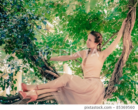 A girl in a delicately pink outfit and high heels sits on a curly tree with green foliage. Blur focus 85369560