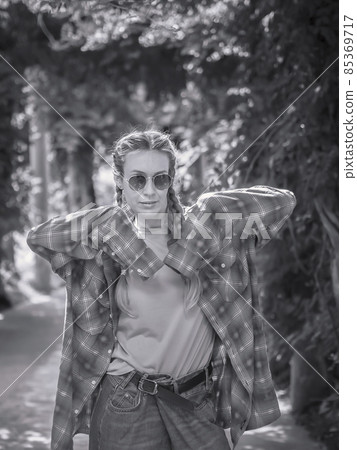 A lady with two braids in a plaid shirt with very long sleeves stands in a pose in the park. Black and white photo. Post-processing. Artistic effect 85369717
