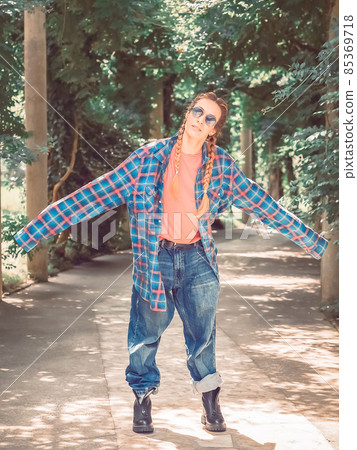 A girl with two braids in a plaid shirt with overly long sleeves and huge jeans stands spreading her arms to the side in the park on a green alley. Post-processing. Artistic effect A girl with two braids in a plaid shirt with overly long sleeves and huge jeans stands spreading her arms to the side in the park on a green alley. Post-processing. Artistic effect 85369718
