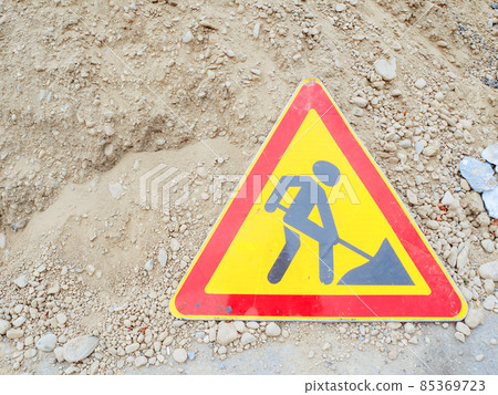 Triangular red-yellow road sign with a picture of a digging man stand near a pile of sand Triangular red-yellow road sign with a picture of a digging man stand near a pile of sand 85369723