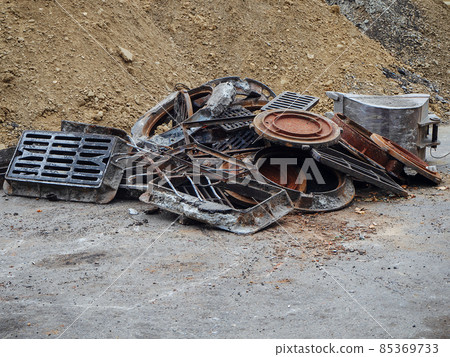 Old rusty metal manhole covers and storm grates piled up near a pile of sand 85369733