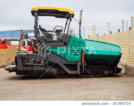 A turquoise empty asphalt paver stands by a construction fence among other machinery on the street A turquoise empty asphalt paver stands by a construction fence among other machinery on the street 85369734