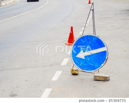 Blue round sign with an arrow and a traffic cone stand on the asphalt road 85369735