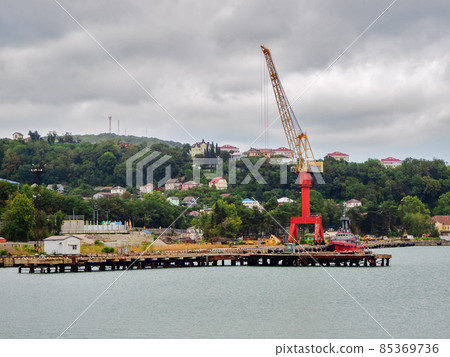 A tall floating crane stands on the pier in the sea near the ship against the backdrop of green hills and houses A tall floating crane stands on the pier in the sea near the ship against the backdrop of green hills and houses 85369736