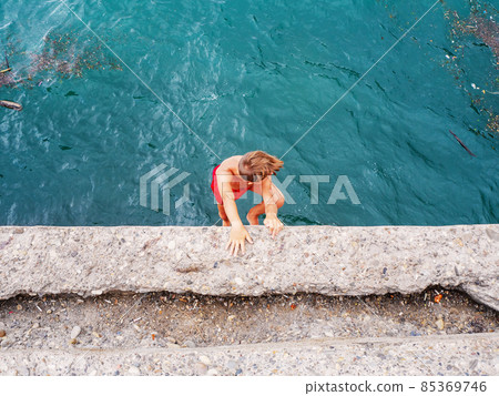 A boy in red swimming trunks descends from a concrete pier into the sea water A boy in red swimming trunks descends from a concrete pier into the sea water 85369746