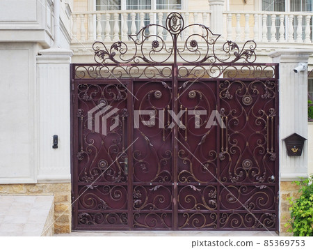 An iron burgundy fence with a forged gilded pattern is located in front of the house with a balcony with concrete balusters An iron burgundy fence with a forged gilded pattern is located in front of the house with a balcony with concrete balusters 85369753