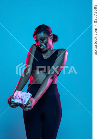 Happy young beautiful dark skinned woman with gift box isolated on blue studio background in neon light. Happy young beautiful dark skinned woman with gift box isolated on blue studio background in neon light. 85373698