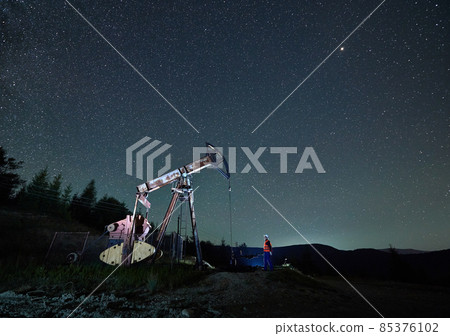 Oil man standing near petroleum pump jack under beautiful night sky with stars. Petroleum engineer controlling work of oil pump rocker-machine at night in oil field. Concept of oil extraction. Oil man standing near petroleum pump jack under beautiful night sky with stars. Petroleum engineer controlling work of oil pump rocker-machine at night in oil field. Concept of oil extraction. 85376102