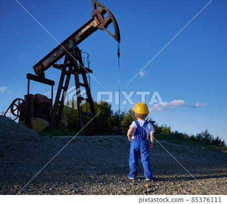 Back view of intelligent child studying business development at oil station with mining platform and drilling rig, boy in yellow helmet and blue uniform for kids looking at machine construction 85376111