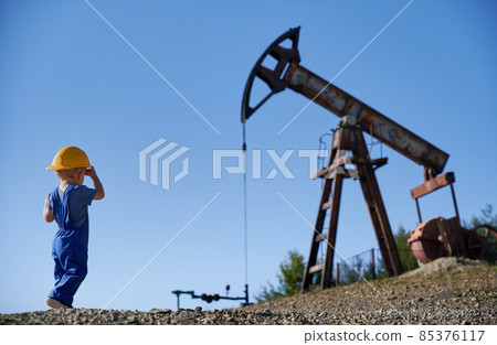 Back view male kid standing on stone surface and looking at working oil drilling rig in petroleum field, on the background of clean blue sky. Back view male kid standing on stone surface and looking at working oil drilling rig in petroleum field, on the background of clean blue sky. 85376117