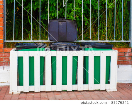 Green trash bins behind a white wooden fence stand outside in front of a fence with green plants behind it 85376241