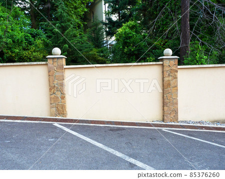 A fence running along a marked asphalt road section and green trees and a fragment of a house behind the fence A fence running along a marked asphalt road section and green trees and a fragment of a house behind the fence 85376260