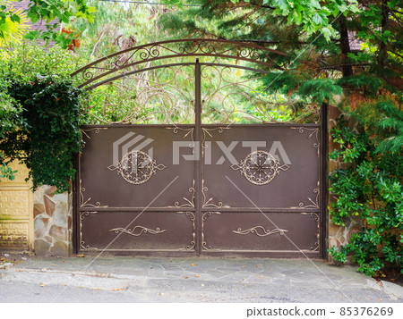 A brown iron gate with a wrought golden pattern and a patterned arch above it and thickets of vegetation next to and behind them A brown iron gate with a wrought golden pattern and a patterned arch above it and thickets of vegetation next to and behind them 85376269